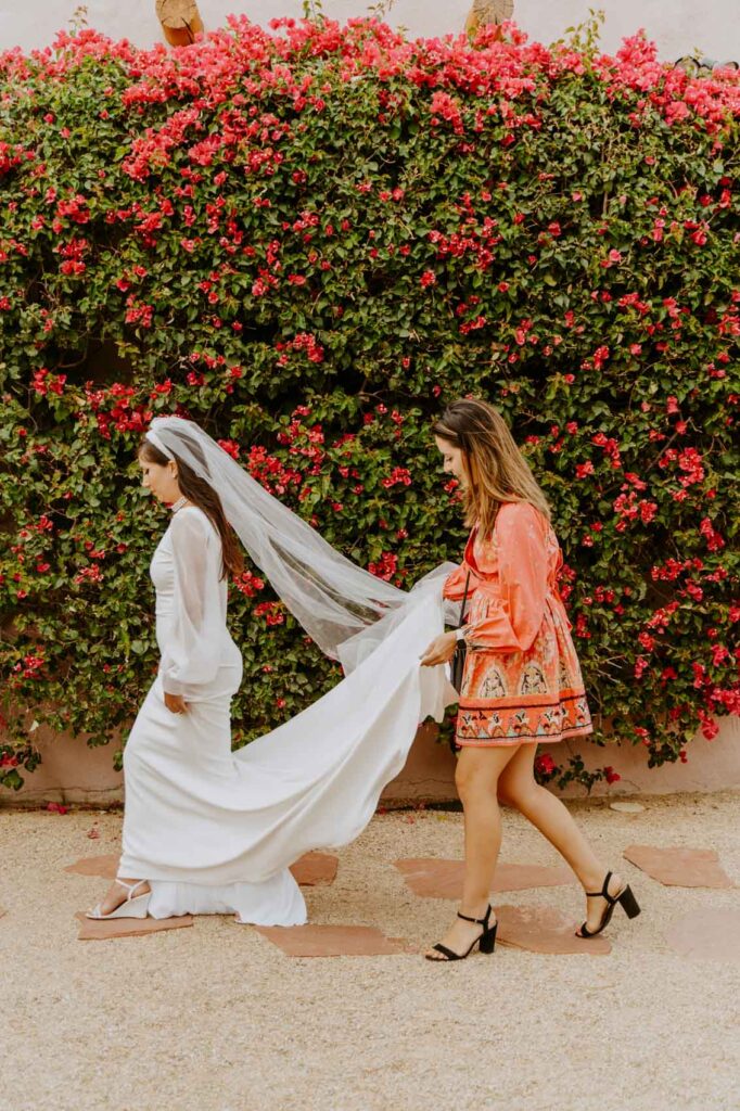 Bridesmaid walking with bride, carrying her train and veil — Desert Wedding Photography by Mattie O'Neill